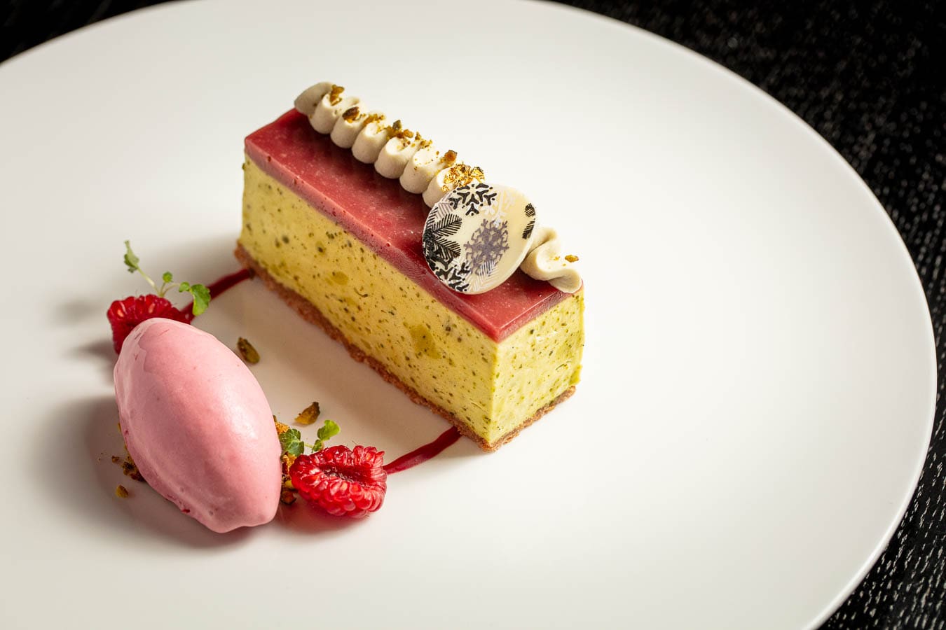A rectangular slice of layered pudding with a pink jelly top, cream, and chocolate décor, served with a scoop of pink sorbet, fresh raspberries, and garnish on a white plate.