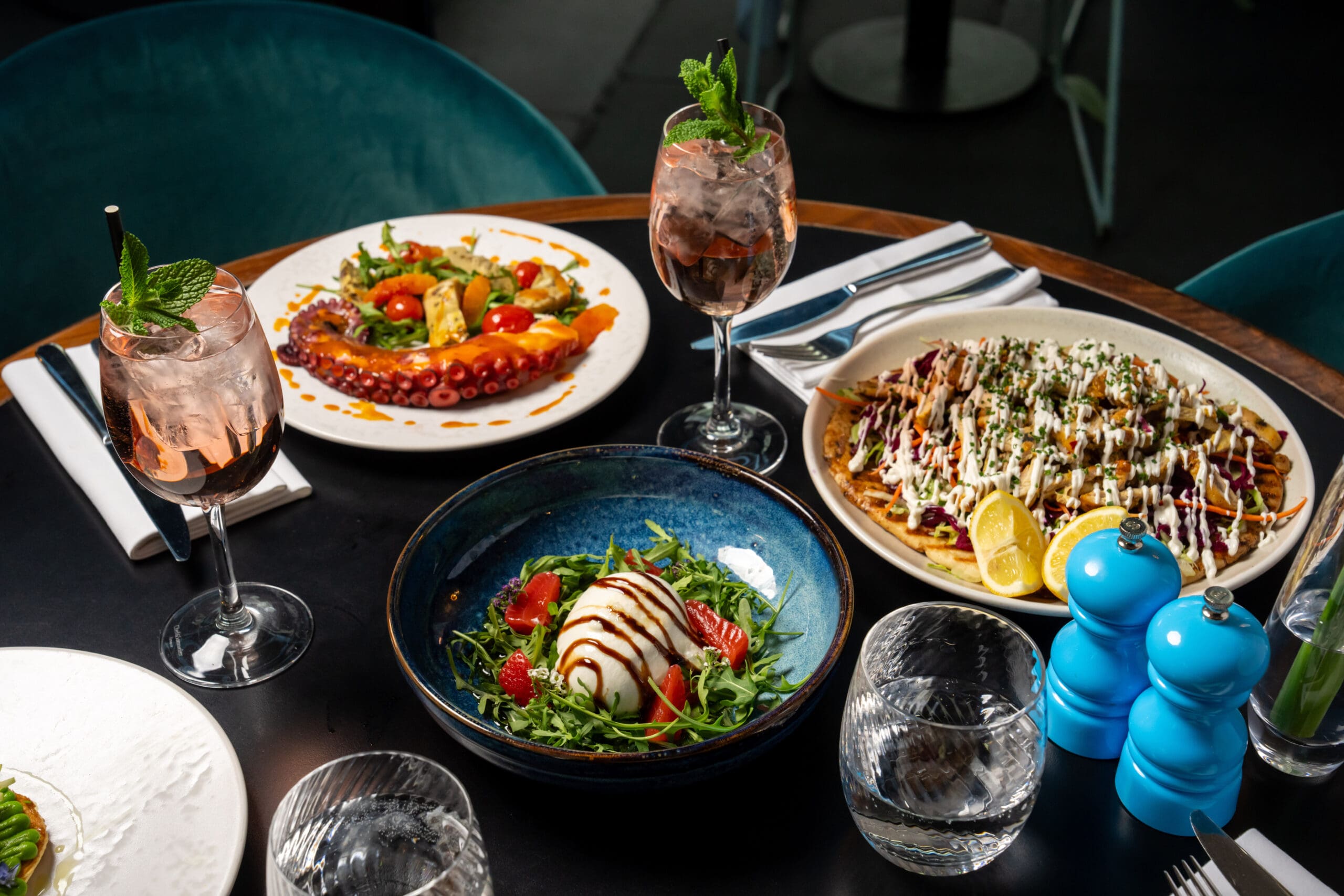 A table laid with gourmet dishes, including grilled octopus, nachos with toppings, a salad with mozzarella and tomatoes, and two glasses of a pink drink garnished with mint, plus blue salt and pepper shakers.