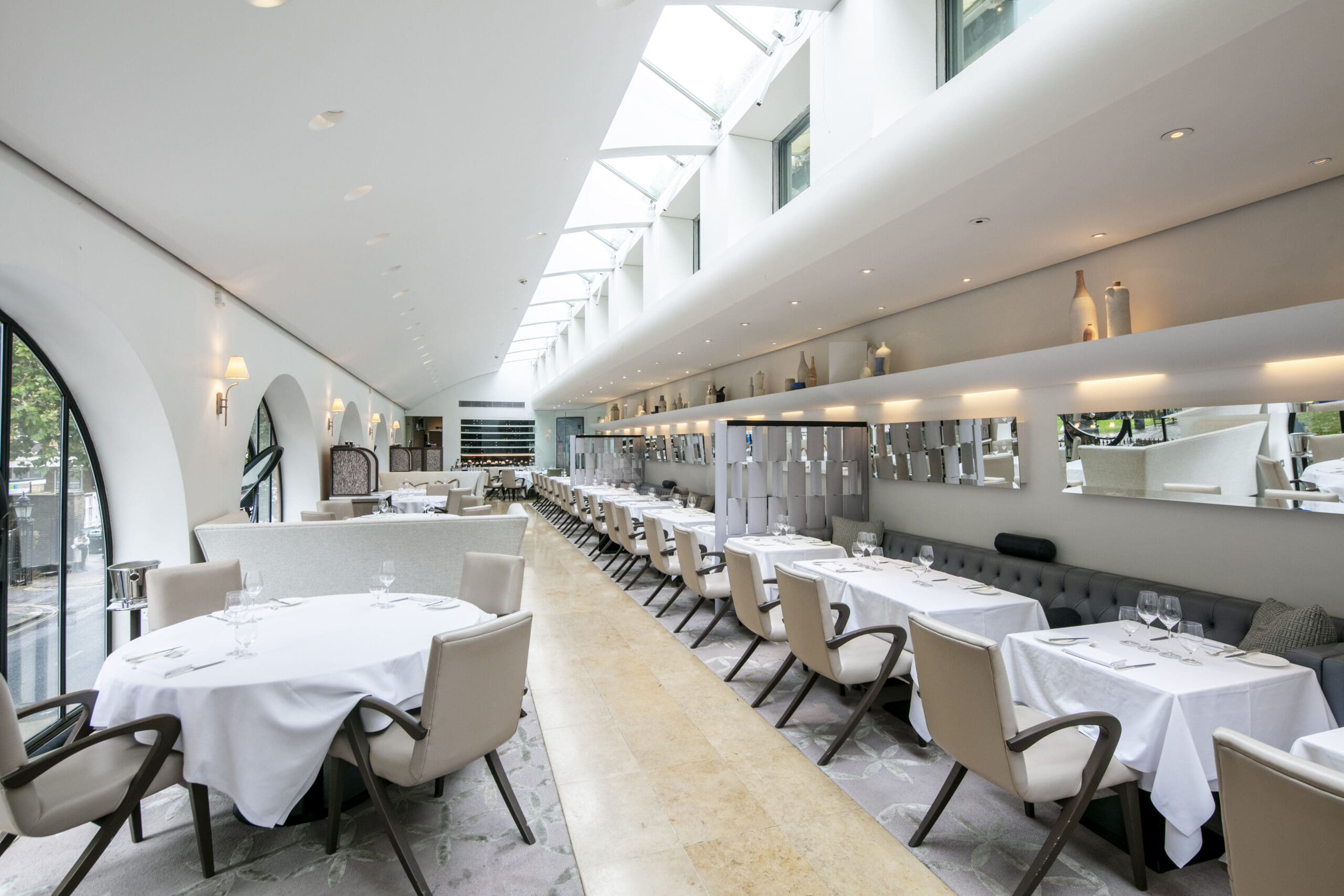 A bright, modern restaurant with skylights, white walls, and neatly arranged tables covered in white cloths. Elegant chairs line both sides, and decorative bottles are displayed on shelves above a large mirror.