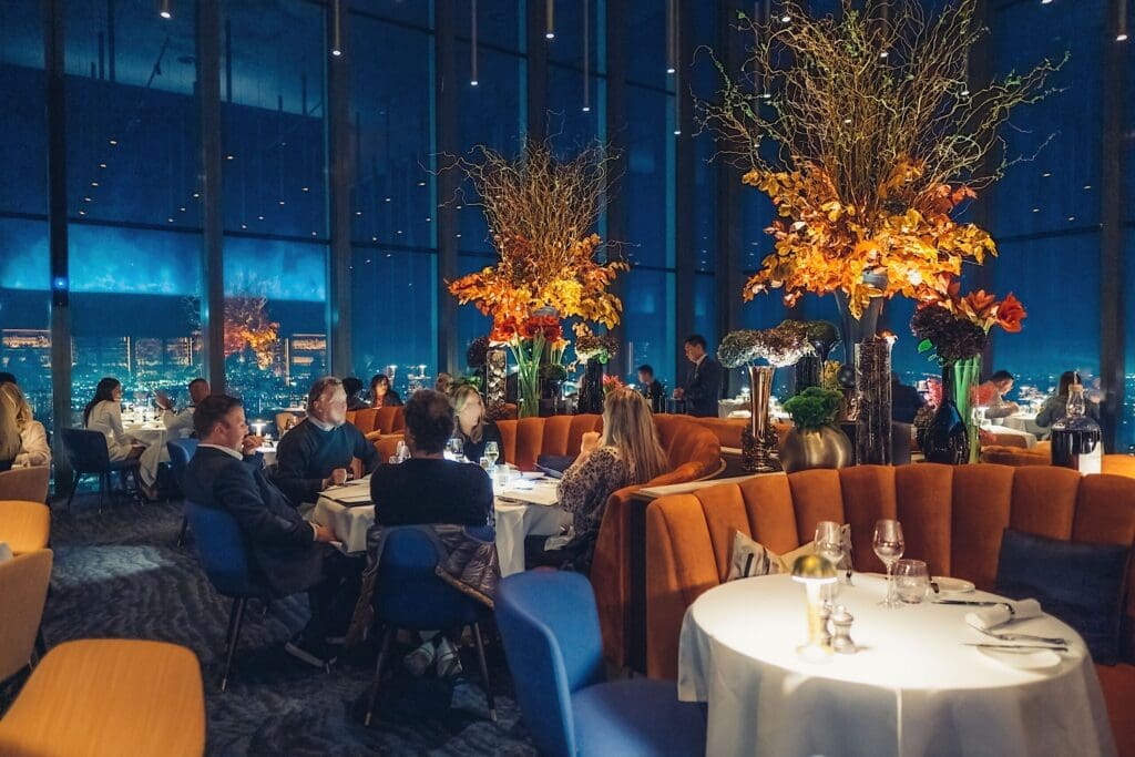 A modern, upmarket restaurant with large windows showing a cityscape at night. Diners sit at round tables with blue and orange chairs; tall autumnal floral arrangements decorate the space, creating a warm, elegant ambience.
