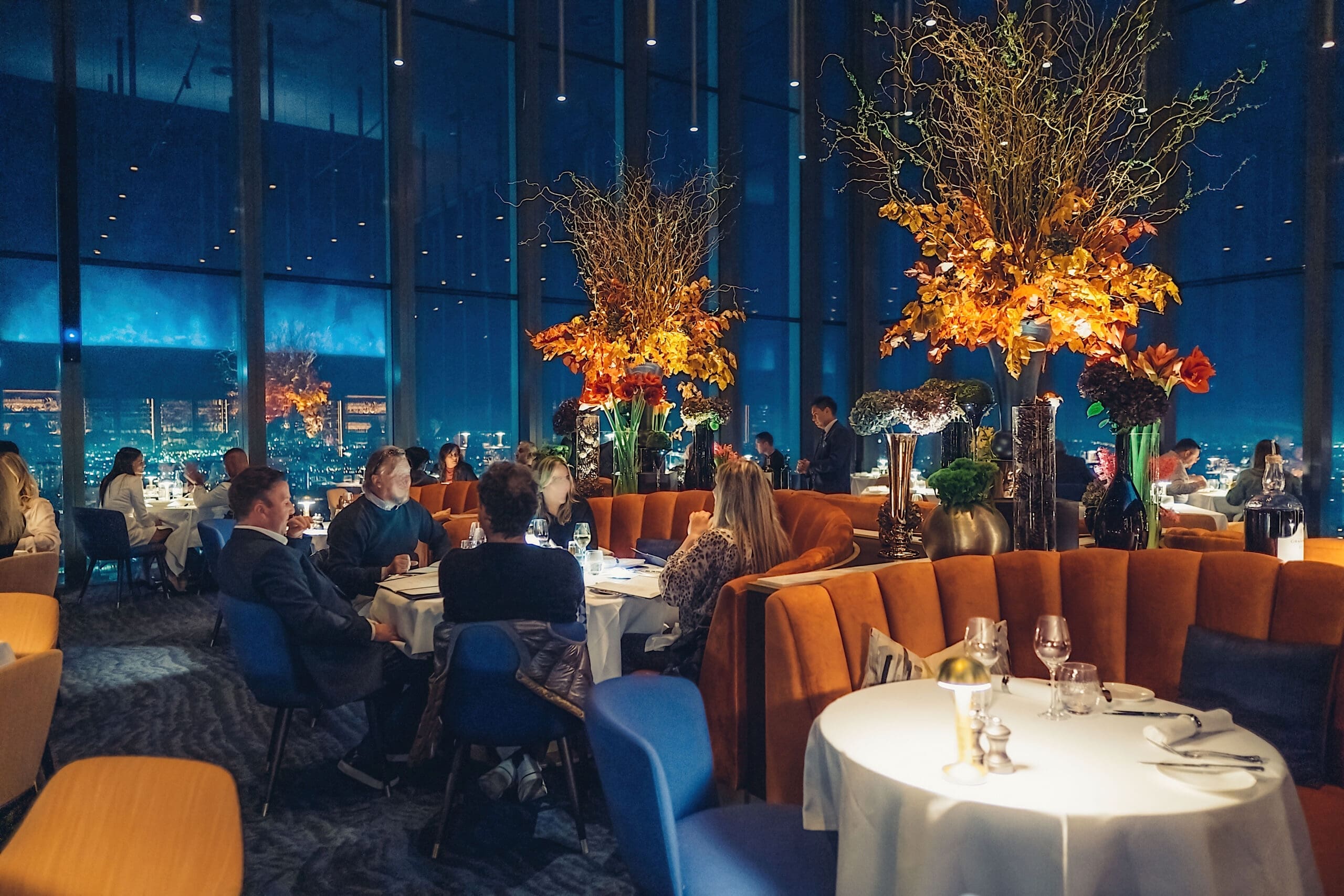 A modern, upmarket restaurant with large windows showing a cityscape at night. Diners sit at round tables with blue and orange chairs; tall autumnal floral arrangements decorate the space, creating a warm, elegant ambience.
